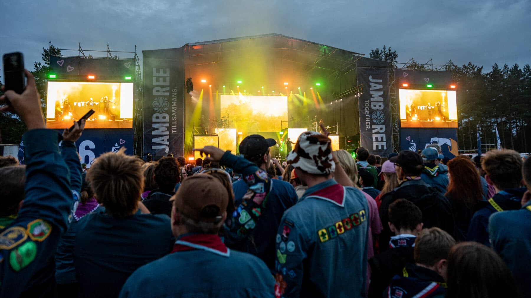 En stor publik, många i scoutuniformer, står framför en scen med färgglada ljus och skärmar. På skärmarna syns artister som uppträder under ett evenemang med texten "Jamboree Tillsammans" på scenens banderoller.